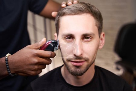Photo for African American barber grooming clients caucasian beard, barber uses special electric razor - Royalty Free Image