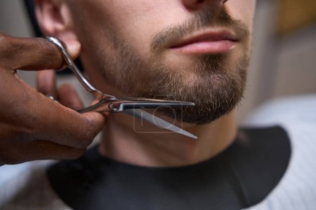 Foto de Barbero afroamericano corta la barba caucásica al cliente, maestro utiliza tijeras especiales - Imagen libre de derechos