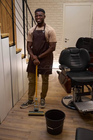 Téléchargez les photos : Un gars souriant dans un tablier de travail nettoie dans un salon de coiffure, il utilise une serpillière et un seau - en image libre de droit
