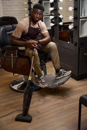 Foto de Peluquero afroamericano en delantal de trabajo descansando en una silla de peluquero, tiene una aspiradora moderna en sus manos - Imagen libre de derechos