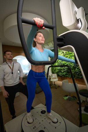 Téléchargez les photos : Attrayant femelle en tenue de fitness faire des exercices soutenus par un entraîneur attentif dans la salle de gym - en image libre de droit