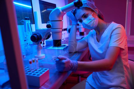 Foto de Mujer joven y cansada en el lugar de trabajo en un laboratorio moderno, sobre una potente mesa de microscopio y tubos de ensayo - Imagen libre de derechos