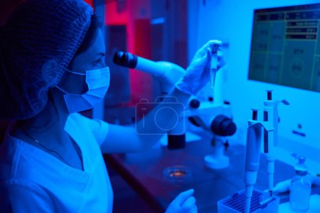 Foto de Mujer sosteniendo un tubo de ensayo con biomaterial en la mano, en el laboratorio potente microscopio - Imagen libre de derechos