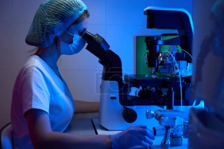 Téléchargez les photos : Embryologue dans des gants de protection et un masque fonctionne avec un biomatériau sous un microscope puissant, équipement moderne dans le laboratoire - en image libre de droit