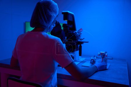Téléchargez les photos : Jeune scientifique dans un masque de protection travaille avec le biomatériau sous un microscope puissant, équipement moderne dans le laboratoire - en image libre de droit