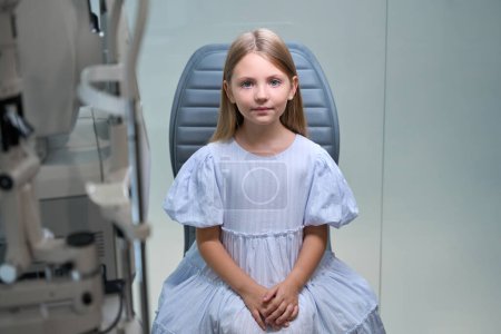 Foto de Jovencita tranquila sentada en silla en el consultorio de oftalmología pediátrica mirando delante de ella - Imagen libre de derechos
