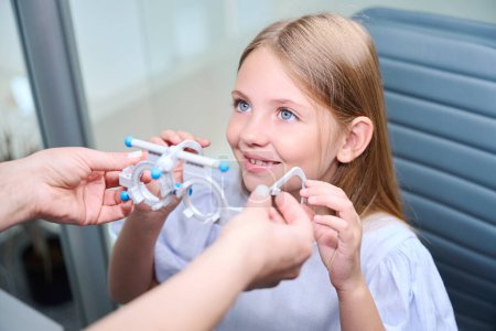 Téléchargez les photos : Optométriste médecin mains tenant cadre d'essai optique devant une petite fille souriante assise dans la chaise d'examen - en image libre de droit