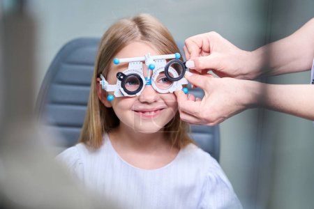 Téléchargez les photos : Mains optométristes insérant des lentilles sphériques dans des cellules de cadre d'essai ophtalmique sur le visage d'une petite fille - en image libre de droit