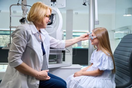 Téléchargez les photos : Ophtalmologiste femelle attachant le cylindre croisé au cadre d'essai optique sur le visage souriant de petite fille - en image libre de droit