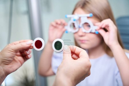 Foto de Manos oftalmólogas sosteniendo filtros rojos y verdes frente al niño con marco de prueba oftálmica en la cara - Imagen libre de derechos