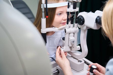 Experienced female oculist looking through slit lamp microscope at little girl eyes
