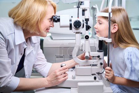 Téléchargez les photos : Souriant amical ophtalmologiste féminine examinant les jeunes yeux du patient à travers le microscope lampe à fente - en image libre de droit