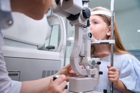 Téléchargez les photos : Optométriste féminine expérimentée examinant les jeunes yeux du patient à travers un microscope à lampe à fente de haute puissance - en image libre de droit