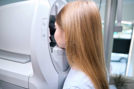 Téléchargez les photos : Vue latérale du patient pédiatrique regardant à l'intérieur de l'autoréfracteur pendant les tests de vision à la clinique - en image libre de droit