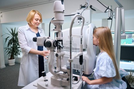 Téléchargez les photos : Ophtalmologiste féminine souriante effectuant une évaluation automatisée de la réfraction oculaire chez l'enfant - en image libre de droit