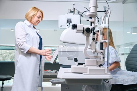 Optometrista femenina focalizada evaluando error refractivo en paciente pediátrica con refractor automatizado