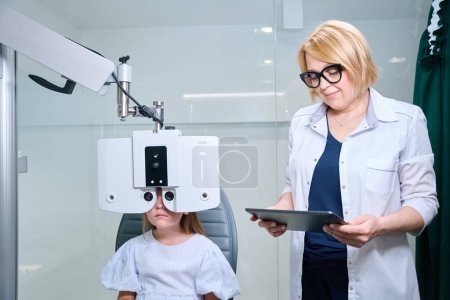Kleines Mädchen sitzt auf Untersuchungsstuhl und blickt durch digitale Phoropterlinsen in Anwesenheit eines Augenarztes