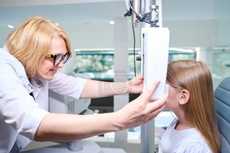 Téléchargez les photos : Orthoptiste féminine souriante plaçant le phoropter numérique devant le visage du patient pédiatrique - en image libre de droit