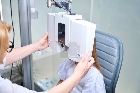 Téléchargez les photos : Expérimenté femelle optométriste positionnement phoropter numérique devant le visage de la petite fille - en image libre de droit