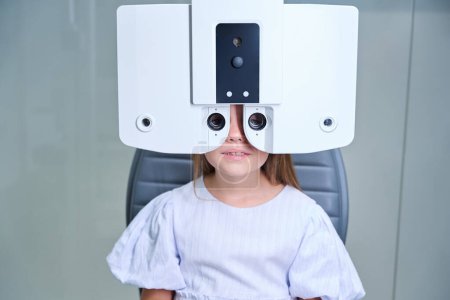 Téléchargez les photos : Portrait d'un patient pédiatrique assis sur une chaise d'examen regardant à travers des lentilles de phoropter numériques - en image libre de droit