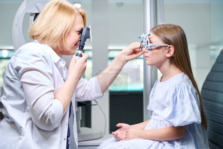 Téléchargez les photos : Vue latérale de l'optométriste regardant à travers le rétinoscope tout en insérant une lentille dans la cellule du cadre d'essai sur le visage du patient pédiatrique - en image libre de droit