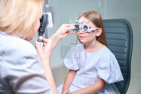 Foto de Ortooptista mirando a través del retinoscopio mientras inserta la lente en la celda del marco de ensayo esférico en la cara de la niña - Imagen libre de derechos