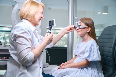 Foto de Optometrista sostiene retinoscopio con tarjeta de texto delante de la cara del niño mientras inserta lente esférica en su celda de marco de prueba - Imagen libre de derechos