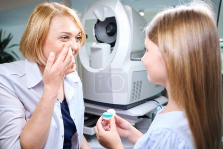 Photo for Qualified female optometrist putting clear contact lens into her right eye in front of child - Royalty Free Image