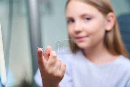 Photo for Child seated on examination chair holding clear contact lens on her fingertip - Royalty Free Image