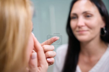 Foto de Primer plano del óptico tirando de su párpado hacia abajo mientras sostiene la lente de contacto en la punta del dedo ante el cliente femenino - Imagen libre de derechos