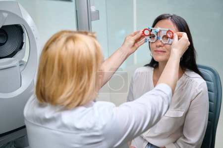 Foto de Profesional oftálmico insertando lentes esféricas en la celda del marco de ensayo frente a los ojos de una mujer adulta - Imagen libre de derechos