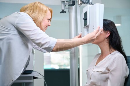 Smiling orthoptist placing electronic phoropter head in front of adult woman eyes