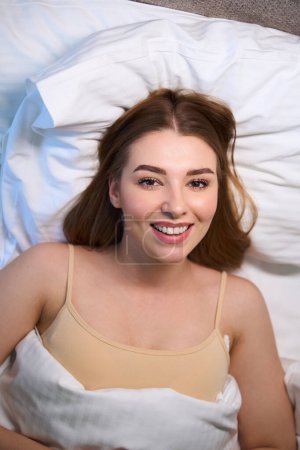 Téléchargez les photos : Vue du dessus de la femme heureuse couverte d'une couverture en soutien-gorge regardant la caméra tout en étant couché sur le lit à l'intérieur - en image libre de droit