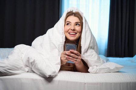 Foto de Mujer feliz con hermosa manicura mirando hacia otro lado mientras sostiene el teléfono celular y se acuesta en la cama en el interior - Imagen libre de derechos