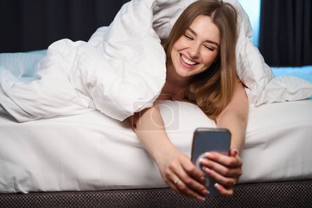 Téléchargez les photos : Heureuse femelle couverte de couverture navigation téléphone cellulaire tout en étant couché sur le lit à l'intérieur - en image libre de droit