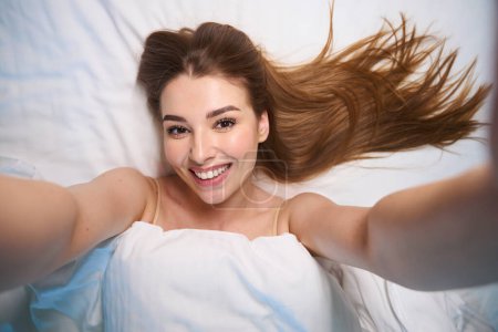 Téléchargez les photos : Selfie de femme heureuse avec les cheveux longs couverts de couverture tout en étant couché sur le lit - en image libre de droit