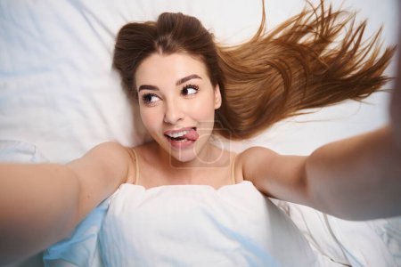 Téléchargez les photos : Selfie de femme heureuse aux cheveux longs recouverts de couvertures faisant des visages tout en étant couché sur le lit - en image libre de droit