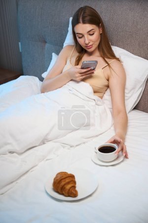 Photo for Caucasian female covered with blanket using mobile while lying on bed with breakfast indoors - Royalty Free Image