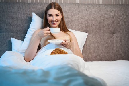 Téléchargez les photos : Femme souriante en soutien-gorge et couverte d'une couverture regardant la caméra tout en buvant du café et couchée sur le lit à l'intérieur - en image libre de droit