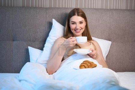 Téléchargez les photos : Femme souriante en soutien-gorge et couverte d'une couverture regardant la caméra tout en buvant du café et couchée sur le lit à l'intérieur - en image libre de droit