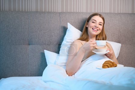 Téléchargez les photos : Femme heureuse en soutien-gorge et couverte d'une couverture regardant la caméra tout en buvant du café et couchée sur le lit à l'intérieur - en image libre de droit