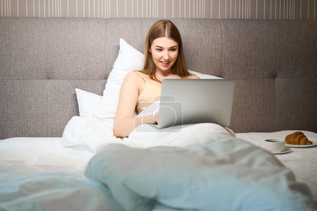 Téléchargez les photos : Femme souriante en soutien-gorge et recouvert d'une couverture à l'aide d'un ordinateur portable tout en étant couché sur le lit à l'intérieur - en image libre de droit