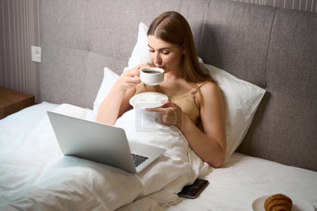 Téléchargez les photos : Belle femme en soutien-gorge et recouvert d'une couverture buvant du café et utilisant un ordinateur portable tout en étant allongé sur le lit à l'intérieur - en image libre de droit