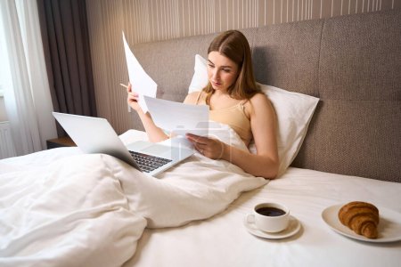 Photo for Concentrated woman in bra and covered with blanket reading notes and using laptop while lying on bed indoors - Royalty Free Image