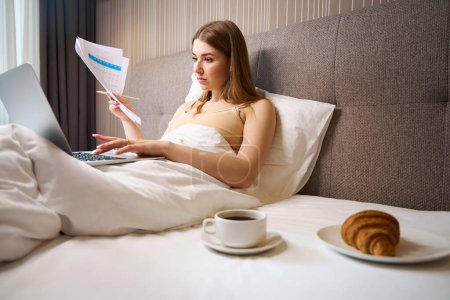 Téléchargez les photos : Belle femme en soutien-gorge et recouvert de couvertures tenant des notes et un ordinateur portable de navigation tout en étant couché sur le lit à l'intérieur - en image libre de droit