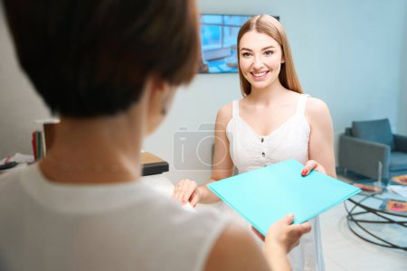 Photo for Two lovely ladies communicate in the hospital lobby, an employee of the clinic passes the patient a folder with documents - Royalty Free Image