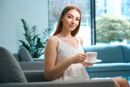 Foto de Mujer joven que se relaja con una taza de té en un sofá acogedor, en el vestíbulo del hospital de diseño ecológico - Imagen libre de derechos