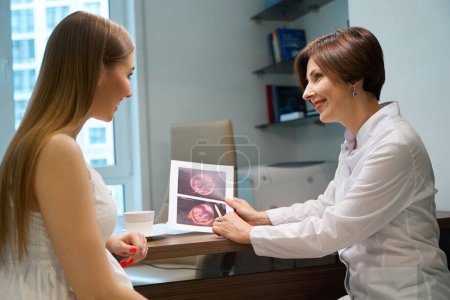Glückliche werdende Mutter untersucht Ultraschallbilder des Fötus gemeinsam mit einem Fruchtbarkeitsspezialisten, Frauen kommunizieren herzlich