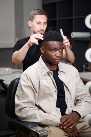 Téléchargez les photos : Coiffeur conseille un Afro-Américain dans un salon de coiffure, le styliste a un outil de travail dans ses mains - en image libre de droit