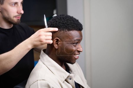 Téléchargez les photos : Coiffeur ramasse une coiffure afro-américaine pour un homme, le styliste a un outil de travail dans ses mains - en image libre de droit